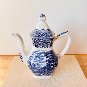 Könnte beinhalten: Eine weiß-blaue Keramik-Teekanne mit Deckel. Die Teekanne hat ein detailliertes blaues Landschaftsdesign und florale Muster. Ausguss und Griff sind weiß. Die Teekanne ist etwa 20 cm hoch.