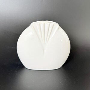 May include: A small, white, ceramic vase with a rounded body and a fan-shaped design on the front. The vase has a smooth, glossy finish and is likely for decorative purposes.