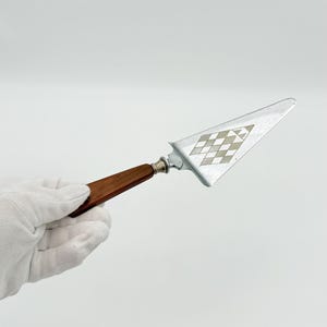 May include: A silver cake server with a brown wooden handle and a decorative diamond pattern on the blade. The server is held by a gloved hand against a white background. A classic serving utensil.