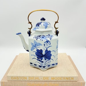May include: A white and blue ceramic teapot with a floral design and a gold handle. The teapot is placed on top of two books. The text "GASTON DIEHL - DIE MODERNEN" is visible on the book.