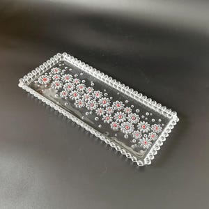 Könnte beinhalten: Ein rechteckiges Tablett aus klarem Glas mit gewelltem Rand. Das Tablett ist mit einem Muster aus weißen Gänseblümchen mit roten Zentren verziert. Das Tablett befindet sich auf einer dunklen Oberfläche.