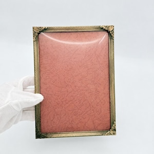 Pode incluir: Uma moldura retangular com acabamento em latão metálico e detalhes decorativos nos cantos. A moldura apresenta uma inserção texturizada na cor rosa seco. As bordas da moldura têm um padrão linear de ranhuras ao longo do perímetro.