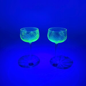 Può includere: Due calici con disegni floreali incisi brillano di una tonalità verde brillante su uno sfondo blu intenso. I calici hanno una coppa rotonda e un gambo lungo e sottile. Il disegno inciso è visibile sulla coppa di ogni calice.