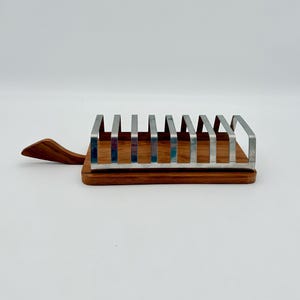 Pode incluir: Um torradeira de madeira com uma alça, com uma série de divisores verticais de aço inoxidável. A base é feita de madeira de tons quentes, e a alça se estende para a esquerda. A torradeira foi projetada para conter várias fatias de torrada.