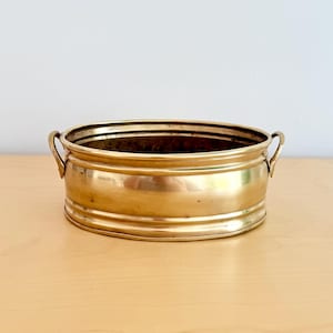 May include: A brass planter, oval in shape, with two small handles. The shiny, golden planter sits on a light-coloured wooden surface. The design features a series of horizontal bands. The planter is approximately 25 cm long.