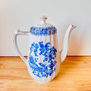 Könnte beinhalten: Weiße Keramik-Kaffeekanne mit blauem Blumenmuster. Die Kanne hat einen Deckel, einen Griff und einen Ausguss, alle mit goldenen Akzenten. Das blaue Muster ist detailliert und bedeckt den Großteil der Oberfläche. Auf einer Holzoberfläche.