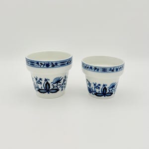 Peut inclure: Deux pots de fleurs en céramique blanche avec des motifs floraux bleus. Les pots ont une bande bleue autour du bord et présentent des motifs floraux bleus détaillés sur le corps. Les pots sont de tailles différentes.