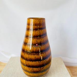 Puede incluir: Un jarrón de cerámica marrón y amarillo con un patrón de rayas. El jarrón tiene un fondo redondeado y una abertura estrecha.