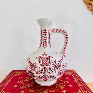 May include: White ceramic pitcher with a handle and floral design in shades of pink, red, and black. The pitcher is decorated with stylized flowers and leaves. The pitcher is sitting on a red surface.