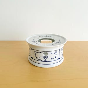 Könnte beinhalten: Ein weißer Stövchen aus Keramik in zylindrischer Form mit einer kreisförmigen Öffnung an der Oberseite. Die Außenseite ist mit einem zarten blauen Blumen- und Rankenmuster verziert. Es verfügt über eine kleine seitliche Öffnung.