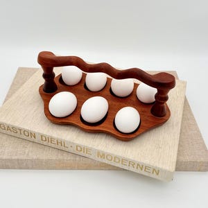Puede incluir: Un soporte de madera oscura para huevos con seis huevos blancos. El soporte tiene un asa ondulada y compartimentos individuales para cada huevo. El soporte descansa sobre una pila de libros. Los libros tienen texto en el lomo.