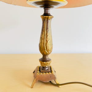Pu&ograve; includere: Una lampada da tavolo vintage con una base dorata e ornata e un paralume color pesca. La base presenta un design dettagliato a forma di foglia e una base quadrata con piedini. La lampada ha un cavo elettrico marrone.