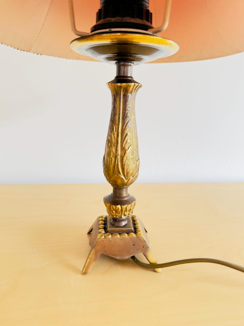 Pu&ograve; includere: Una lampada da tavolo vintage con una base dorata e ornata e un paralume color pesca. La base presenta un design dettagliato a forma di foglia e una base quadrata con piedini. La lampada ha un cavo elettrico marrone.