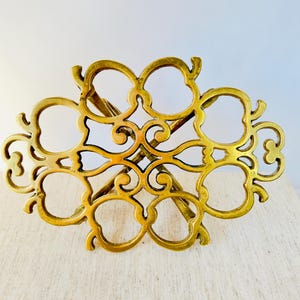 Könnte beinhalten: Ein goldfarbener Metall-Topfuntersetzer mit einem komplizierten, verzierten Design. Der Topfuntersetzer ist blumenförmig mit mehreren abgerundeten Blütenblättern.