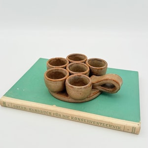 Könnte beinhalten: Ein Set aus sechs kleinen, rustikalen Keramikbechern, angeordnet auf einem passenden Tablett mit Griff. Die Becher und das Tablett sind braun gesprenkelt und stehen auf einem grünen Buch.