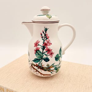 Puede incluir: Cafetera de cerámica blanca con tapa, con un diseño floral con flores rosas, hojas verdes y raíces marrones. La cafetera tiene un asa y un pico. El diseño se repite en la tapa.