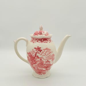 Puede incluir: Una tetera de cerámica blanca con un diseño de paisaje rojo. La tetera tiene un asa curva, un pico largo y una tapa con un pequeño adorno. El diseño del paisaje presenta árboles, edificios y montañas.