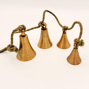 May include: Four bronze-colored bells with a ridged texture are suspended from a twisted, olive-green rope. The bells are of varying sizes and are knotted to the rope. The background is a plain white.