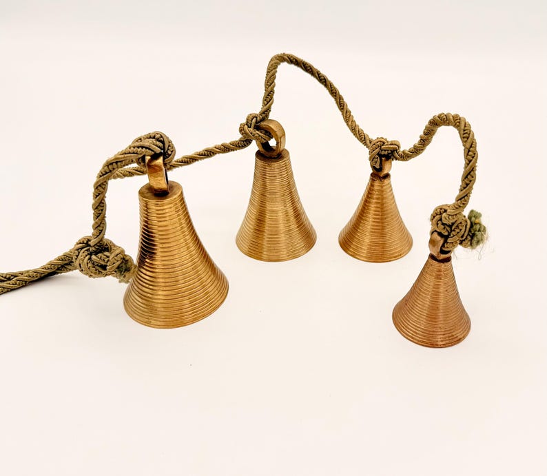 May include: Four bronze-colored bells with a ridged texture are suspended from a twisted, olive-green rope. The bells are of varying sizes and are knotted to the rope. The background is a plain white.