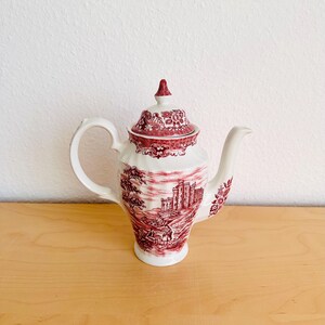Può includere: Una teiera in ceramica bianca e rossa con coperchio, manico e beccuccio. La teiera presenta una dettagliata scena paesaggistica rossa e motivi floreali. La teiera è posizionata su una superficie di legno.