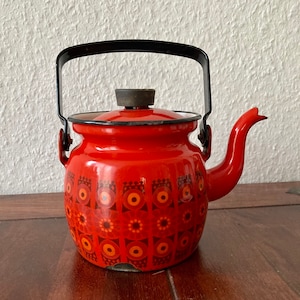 May include: A vintage red enamel teapot with a black handle and lid. The teapot features a decorative pattern of orange and brown floral designs. The spout is curved, and the teapot sits on a wooden surface.