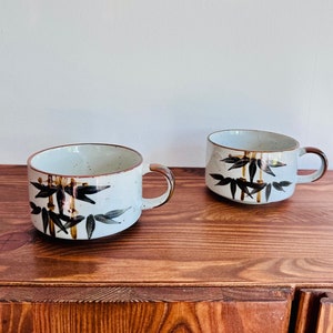 May include: Two ceramic mugs with a white speckled finish and brown trim. Each mug features a bamboo design in black and brown. The mugs have a rounded shape with a handle and are displayed on a wooden surface.