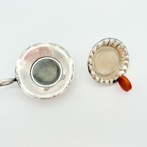 Puede incluir: Dos coladores de té antiguos de plata. Uno es redondo con un asa y un filtro de malla. El otro es más pequeño, con un borde decorativo y un asa de madera. Ambos están sobre un fondo blanco.