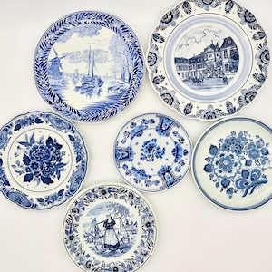 May include: A collection of six ceramic plates featuring blue and white designs. The plates display various illustrations including a windmill, a large building, floral patterns, and a person carrying baskets, all with intricate blue decorative borders.