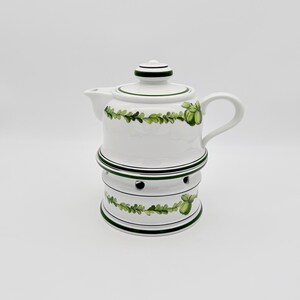 Può includere: Teiera in ceramica bianca con scaldavivande abbinato. La teiera ha coperchio e manico ed è decorata con motivi a foglie e bacche verdi. Lo scaldavivande ha fori di ventilazione ed è anch'esso decorato con motivi a foglie e bacche verdi.