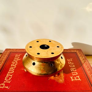 Könnte beinhalten: Ein kleiner, antiker Räuchergefäß aus Messing mit einem runden, perforierten Deckel und einem bauchigen Körper. Der Brenner steht auf drei kleinen Füßen und hat mehrere kreisförmige Öffnungen. Er liegt auf einem roten Buch mit goldener Schrift, das "PICTURESQUE EUROPE" lautet.