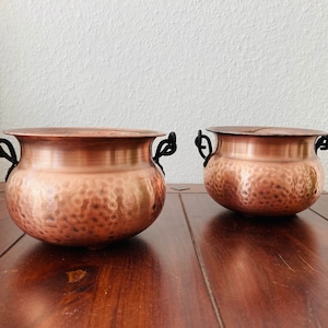 Op de afbeelding: Twee gehamerde koperen potten met zwarte ijzeren handvatten. De potten staan op een houten oppervlak.