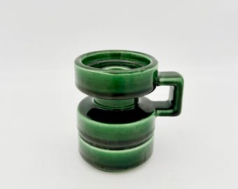 May include: A dark green ceramic mug with a glossy finish, featuring a stacked cylindrical body design and a square-shaped handle. The mug has a distinct tiered appearance with a wide rim and a smooth, reflective surface throughout the piece.