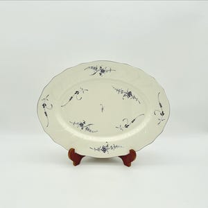May include: An oval, off-white serving platter with a scalloped edge and dark blue floral designs. The platter is supported by a small, brown wooden stand. The floral pattern is repeated around the edge of the platter.