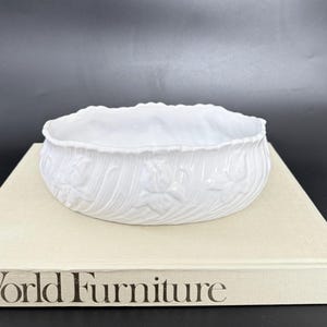 Peut inclure: Un bol ovale en céramique blanche avec un motif floral texturé sur l'extérieur. Le bol est posé sur un livre avec les mots "World Furniture" imprimés sur la couverture. Le bol a un bord festonné et est posé sur un fond sombre.