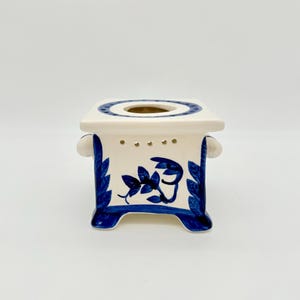 Könnte beinhalten: Ein quadratischer Keramik-Ölwärmer mit weißer Basis und handgemalten blauen Blumen- und Blattmotiven. Die Oberseite hat eine kreisförmige Öffnung, und die Seiten verfügen über kleine Belüftungslöcher über den Mustern sowie vier Standfüße.