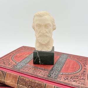 Könnte beinhalten: Eine kleine, cremefarbene Büste eines bärtigen Mannes auf einem schwarzen Marmorsockel. Die Büste steht auf einem Stapel antiker Bücher mit roten Einbänden und aufwendigen Gold- und Silberdetails. Die Bücher haben abgenutzte Einbände.