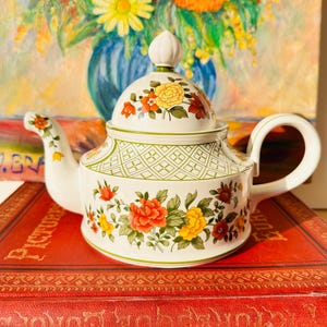 May include: A white ceramic teapot with a floral design featuring red, yellow, and orange flowers. The teapot has a curved handle and a lid with a small knob. The teapot is sitting on a red book.