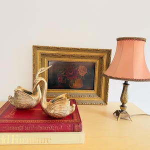 Pu&ograve; includere: Una natura morta con due statuette di cigni dorati, un dipinto floreale incorniciato e una lampada color pesca con base decorativa. Gli oggetti sono disposti su un tavolo con due libri.