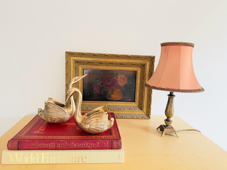 Pu&ograve; includere: Una natura morta con due statuette di cigni dorati, un dipinto floreale incorniciato e una lampada color pesca con base decorativa. Gli oggetti sono disposti su un tavolo con due libri.