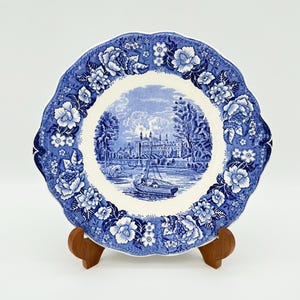 May include: A blue and white decorative plate with a floral border and a central landscape scene. The plate features a scalloped edge and is displayed on a wooden stand. The landscape scene depicts a building, trees, and a boat on the water.