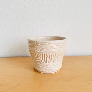Könnte beinhalten: Ein beigefarbener Keramik-Pflanztopf mit einem strukturierten, holzähnlichen Design. Der Topf hat eine zylindrische Form mit einem leicht ausgestellten Rand. Das strukturierte Muster umschließt den Körper des Topfes und sorgt für visuelles Interesse. Der Topf steht auf einer Holzoberfläche.