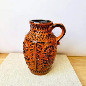 Könnte beinhalten: Ein orangefarbener Keramikkrug mit Henkel, mit strukturierter Oberfläche und aufwendigen geprägten Mustern. Der Krug hat einen dunklen Rand und Henkel und steht auf einer hellen Oberfläche.