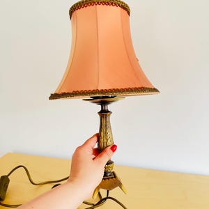 Pu&ograve; includere: Una lampada da tavolo vintage con un paralume color pesca e una base decorativa in bronzo. Il paralume &egrave; rifinito con dettagli dorati e marroni. La lampada &egrave; collegata e pronta all'uso. Un pezzo classico per l'arredamento della casa.