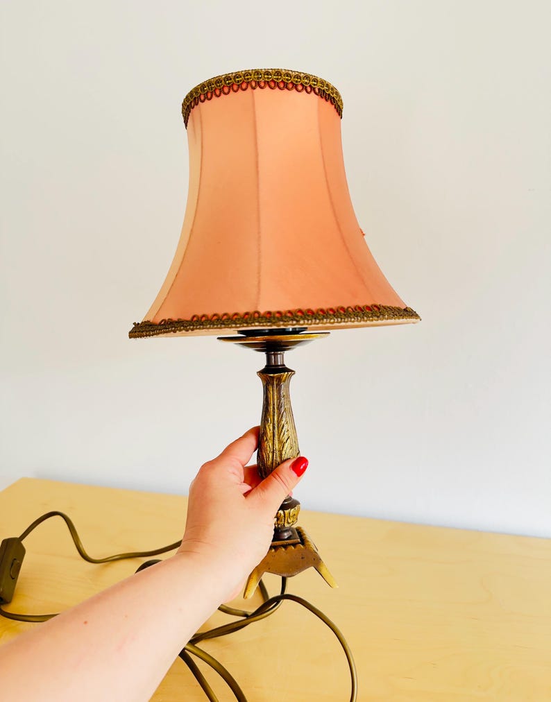 Pu&ograve; includere: Una lampada da tavolo vintage con un paralume color pesca e una base decorativa in bronzo. Il paralume &egrave; rifinito con dettagli dorati e marroni. La lampada &egrave; collegata e pronta all'uso. Un pezzo classico per l'arredamento della casa.