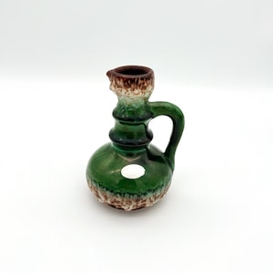 May include: A small, green ceramic pitcher with a handle and spout. The pitcher has a bulbous base and a textured, brown and white rim. A white sticker is on the front of the pitcher. The pitcher is likely vintage.