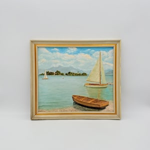 Op de afbeelding: Vierkant ingelijst olieverfschilderij van een meerlandschap met zeilboten en een kleine houten boot. Het water is lichtblauw-groen, met een lichtblauwe lucht en witte wolken. De lijst is beige.