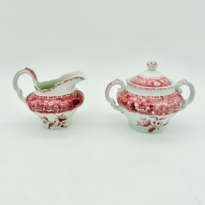 May include: A white ceramic creamer and a lidded sugar bowl featuring a red floral transferware pattern. Both pieces have ornate handles and a scalloped rim on the creamer, with intricate botanical designs covering the lower sections of each.