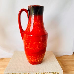 Puede incluir: Una jarra alta de cerámica roja con borde y asa negros. La jarra tiene una superficie texturizada y está colocada sobre dos libros. Los libros tienen los títulos "GASTON DIEHL - DIE MODERNEN" y "World Furniture".