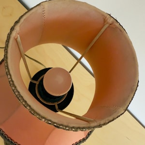 Pu&ograve; includere: Un paralume vintage con interno in tessuto color pesca e finiture decorative. Il paralume ha una forma arrotondata ed &egrave; sostenuto da un telaio in metallo. La base della lampada &egrave; nera.