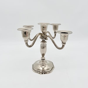 May include: A silver-toned metal candelabra featuring a central pillar and four curved arms extending outward. Each of the five arms terminates in a circular candle cup with a wide, flat rim. The base is circular with a scalloped edge design.